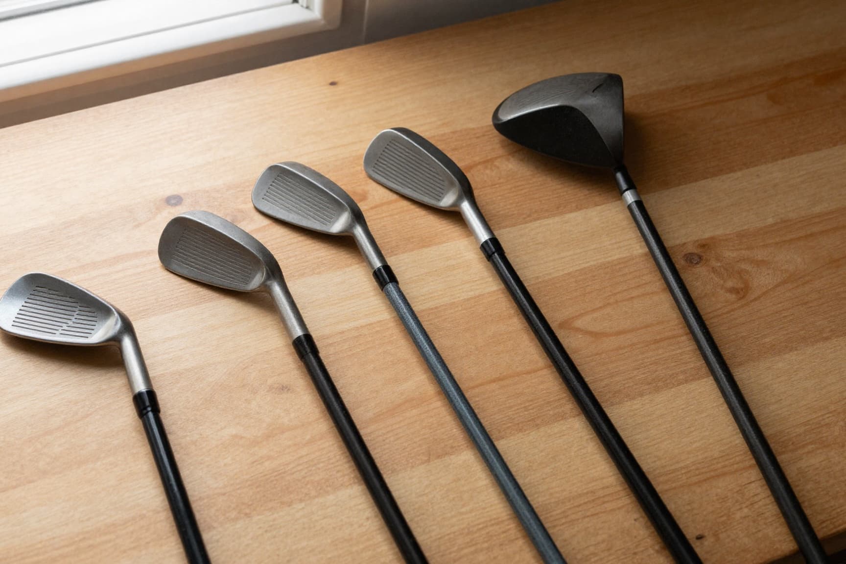 Several pre-owned golf clubs arranged on warm wood surface in soft window light
